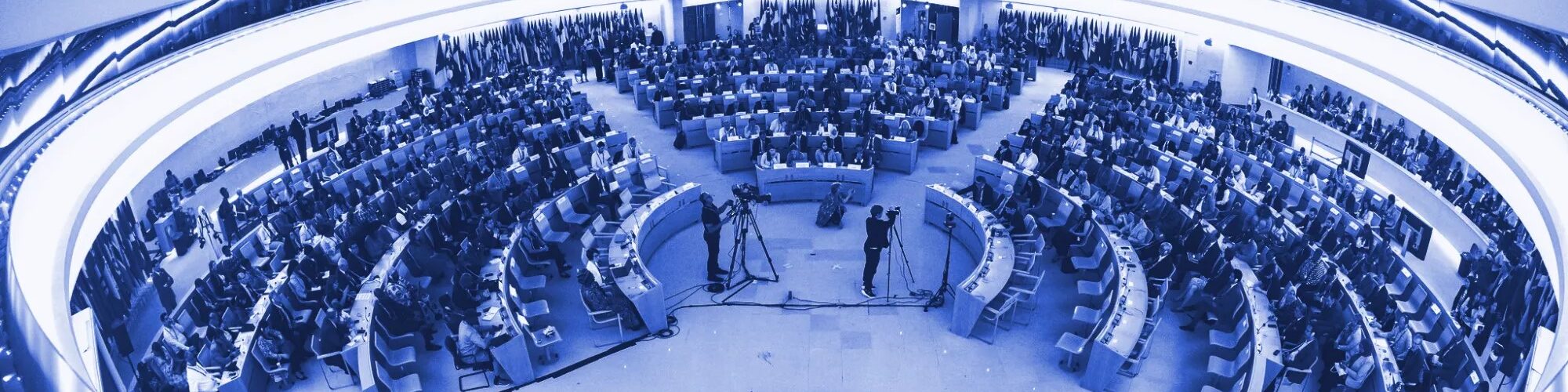 Imagem da sala de conferências da OIT. Lugares dispostos em circulos e onde estão sentados os representantes dos Estados Membros. A sala está com luz ambiente luz.