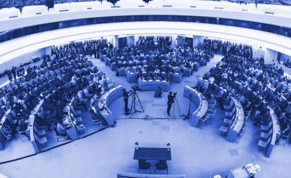 Imagem da sala de conferências da OIT. Lugares dispostos em circulos e onde estão sentados os representantes dos Estados Membros. A sala está com luz ambiente luz.